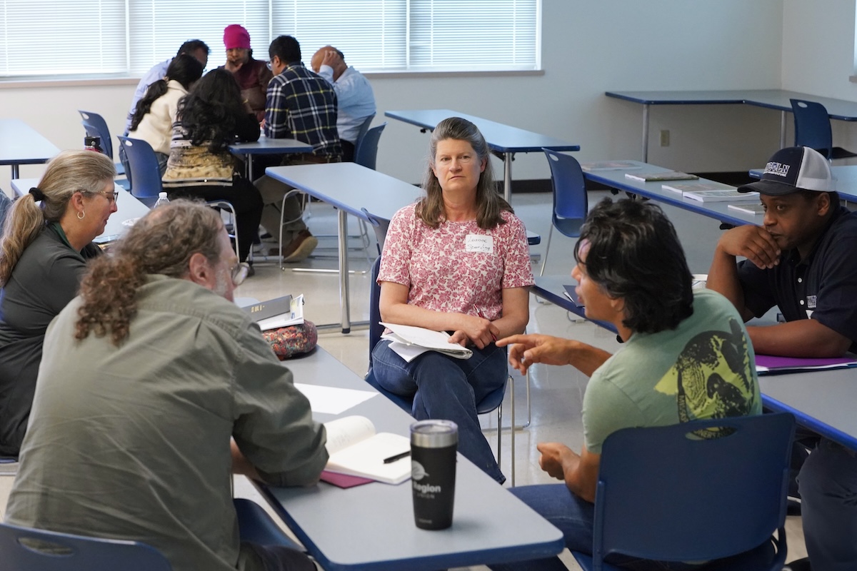 Five people sitting closely at tables speak with each other. 