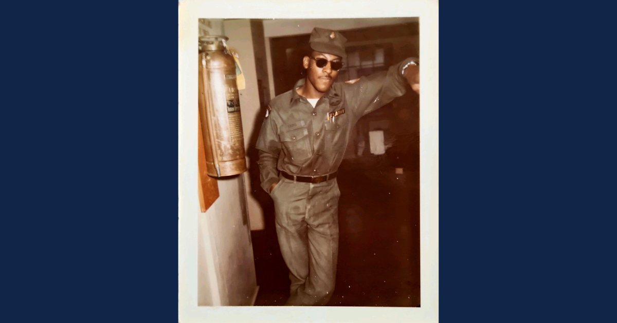 A young African American man in a vintage U.S. Army uniform leans casually against a wall indoors, wearing sunglasses and a confident expression. A brass fire extinguisher is mounted on the wall beside him. The photo has a nostalgic, mid-20th-century aesthetic.